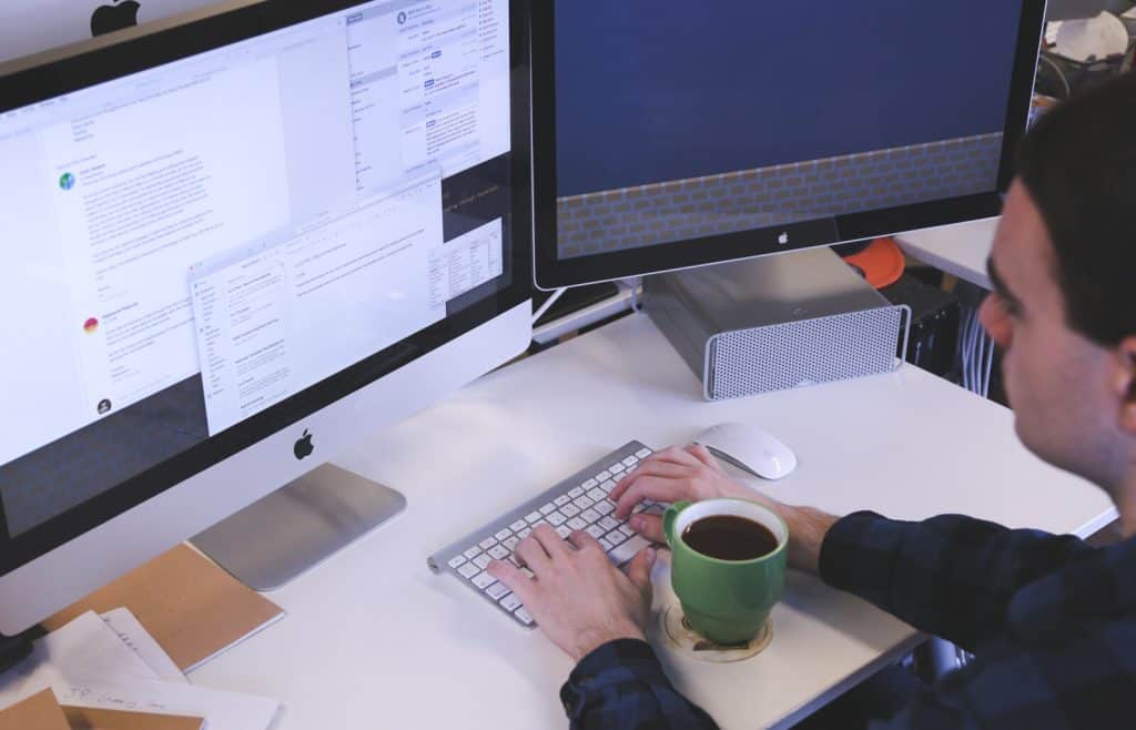 A man sitting at a desk with two monitors and a cup of coffee.