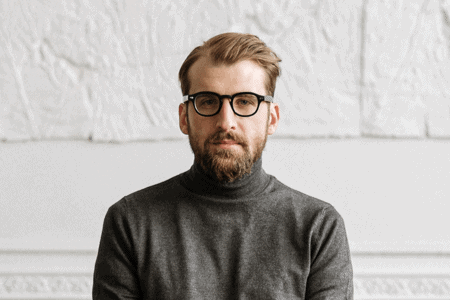 A bearded person wearing glasses and a gray turtleneck stands against a textured white wall.