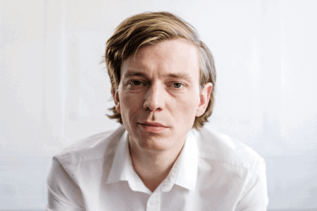 A person with short hair wearing a white shirt, looking directly at the camera with a neutral expression against a plain background.